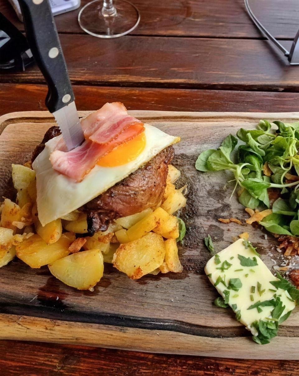 Steak topped with fried egg and bacon on a wooden board, with roast potatoes and salad